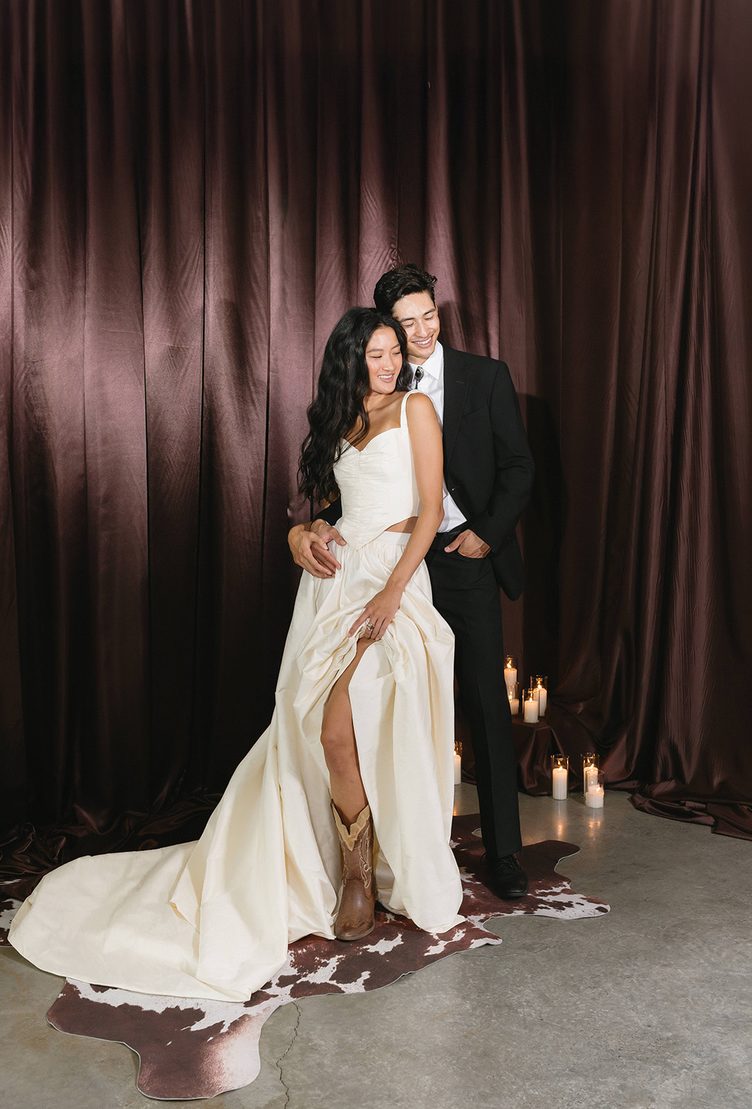 Bride in an ivory gown and cowboy boots with a groom in a black suit, before brown satin drapes
