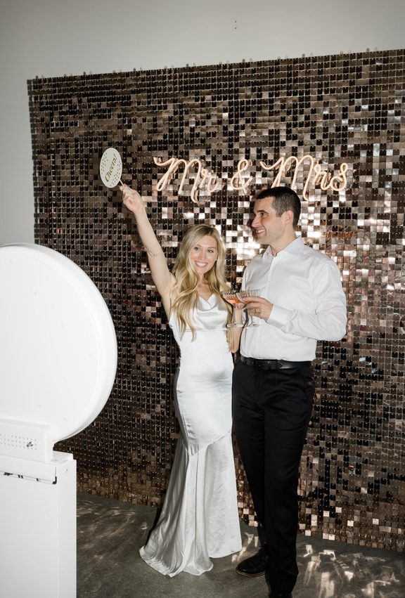Couple at a sequin wall with a lit "Mr & Mrs" sign beside a white photo booth