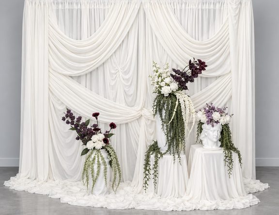 White draped backdrop with white, burgundy and purple floral arrangements and trailing greenery, one in a sculpted bust vase